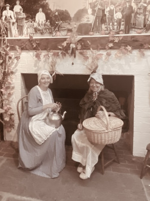 Two women in pioneer costumes sitting in front of a fireplace