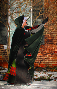 woman in hooded black cloke holding arm up in front of old brick building