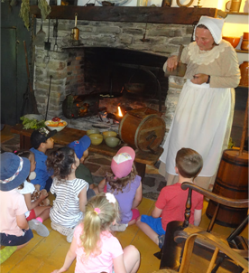 woman in front of fireplace dressed in 19th centry clothing standing in front of children who are sitting